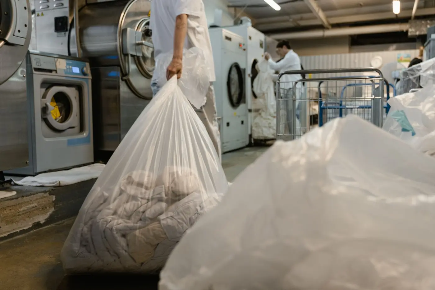 A worker carrying a large clear bag of laundry past industrial washers